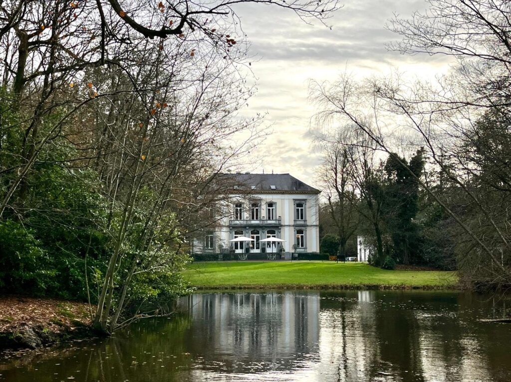 We zien een grote witte villa omgeven door een meer en natuur